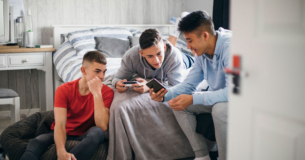 Three teenage boys show each other content on their smartphones in a bedroom. 