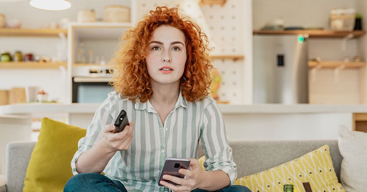 A woman watches TV on her couch with a remote and mobile phone