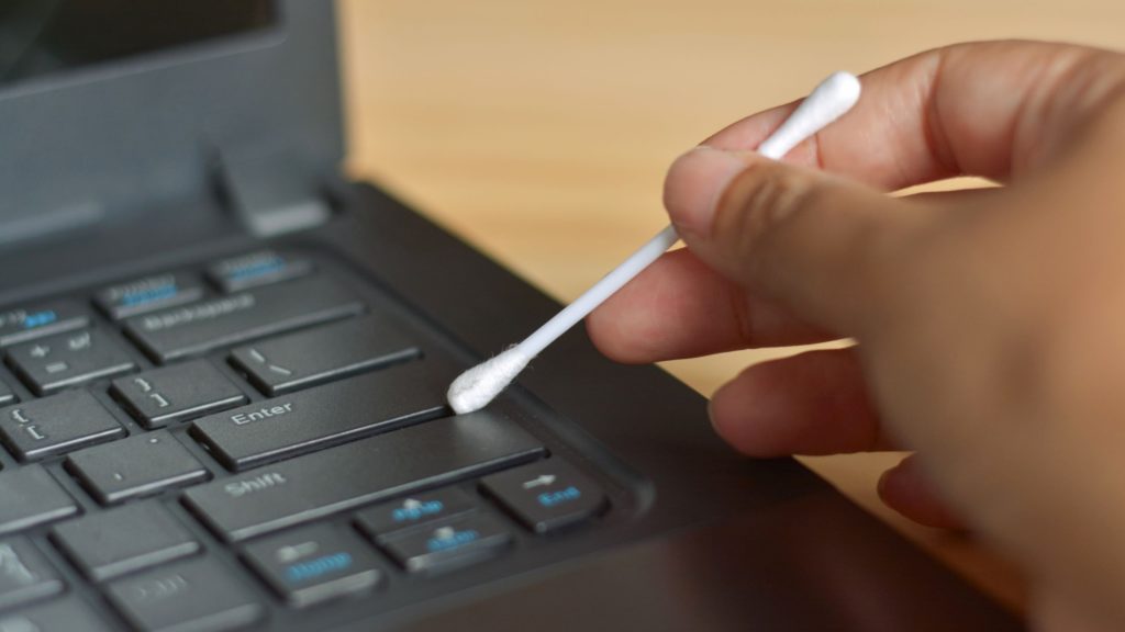 A person uses a q-tip to clean between the keys of a laptop keyword, cleaning your computer. 