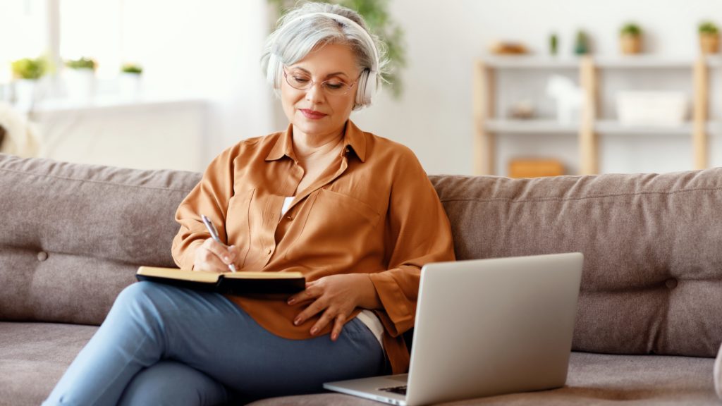 A woman listens to an online business course from her couch and takes notes.