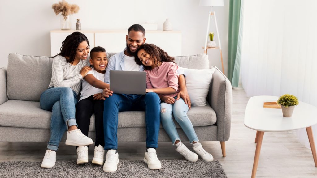 A family watches a video together on a laptop as they sit on the couch. 