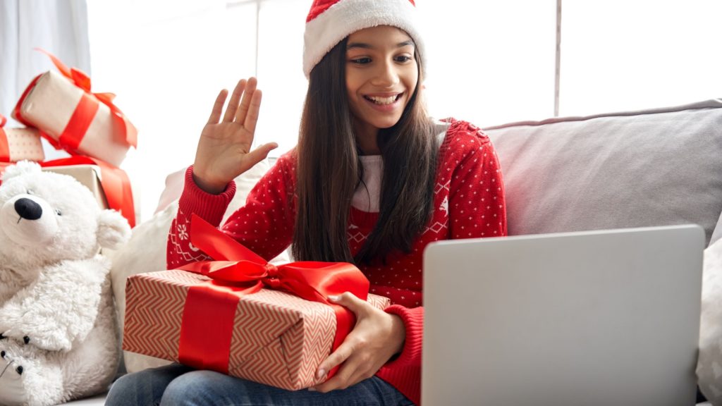 A young woman waves hello on a video chat as she opens up a holiday gift. 