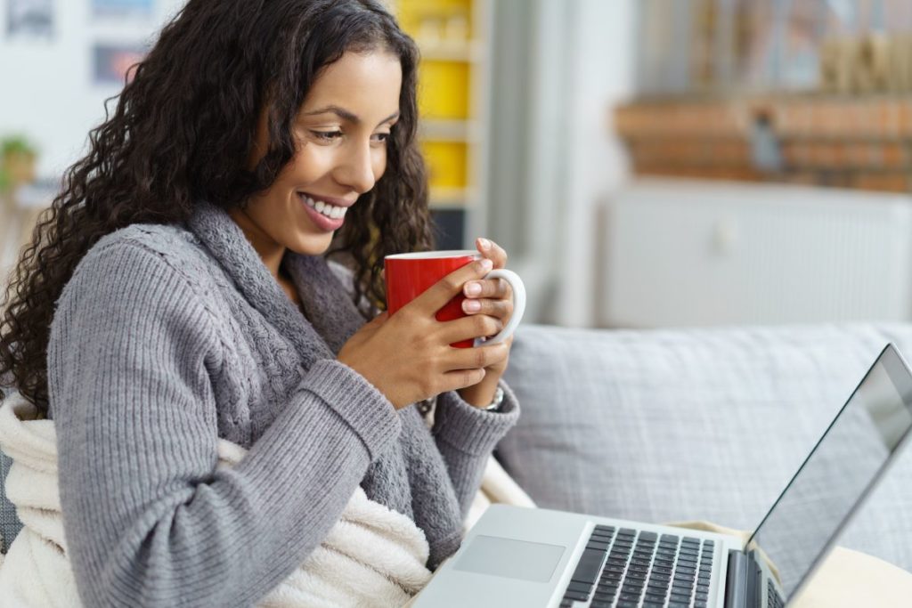 A woman watches a show on her laptop on the couch as she drinks a warm cup of tea.
